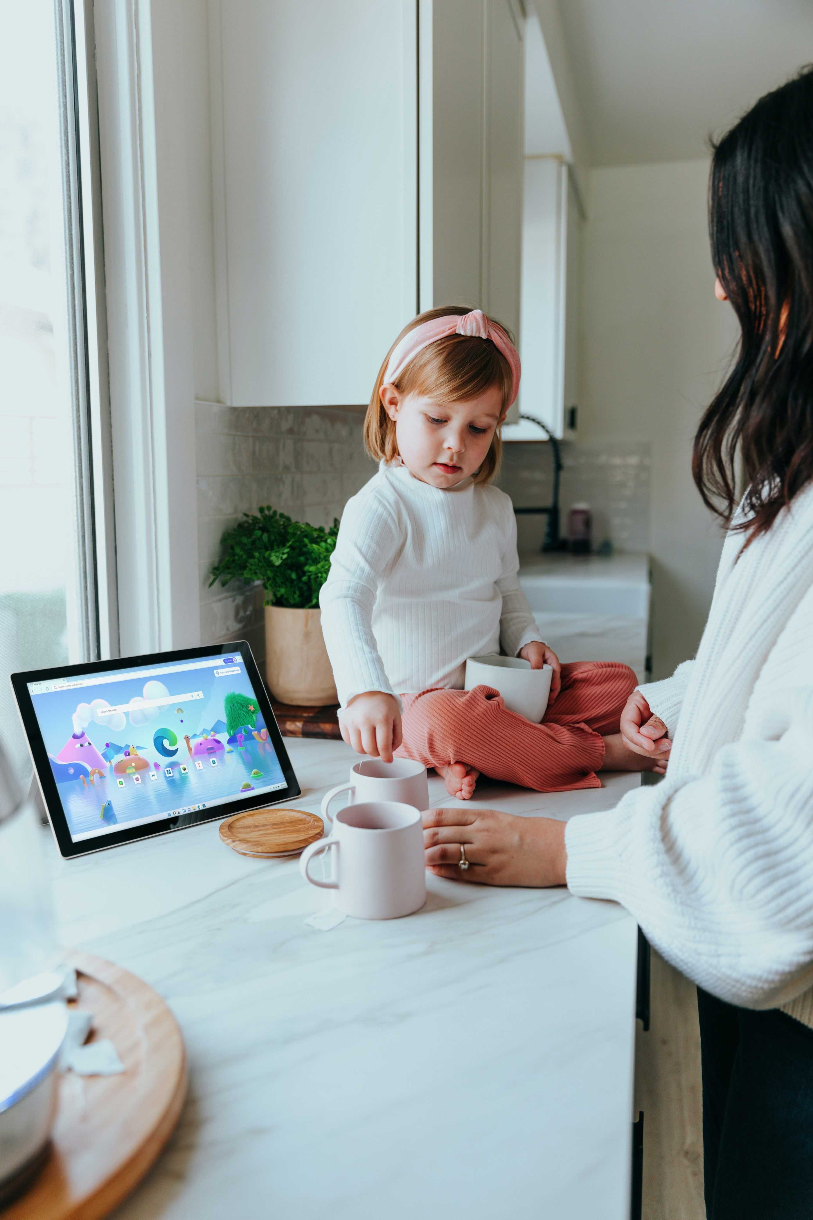 Lille pige sider på køkkenbordet og ser noget på en tablet mens moren står ved siden af